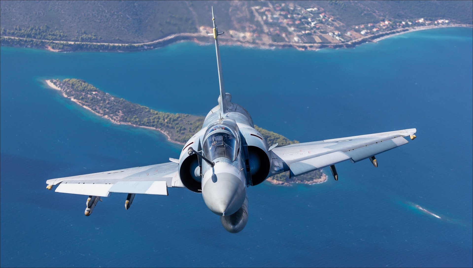 HD desktop wallpaper featuring a Dassault Mirage 2000 military jet soaring above a coastal landscape with clear blue water and islands below.