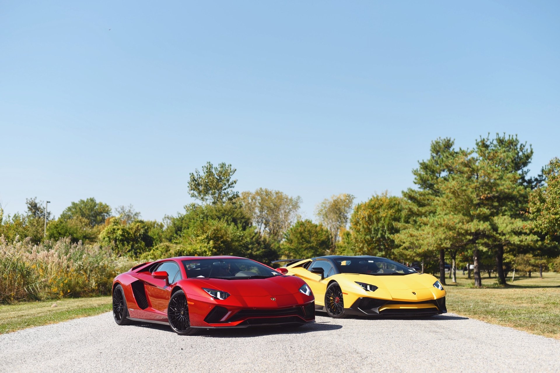 Two Lamborghini Aventador sports cars, red and yellow, parked on a sunlit road by trees — 4K Ultra HD PC desktop wallpaper background.
