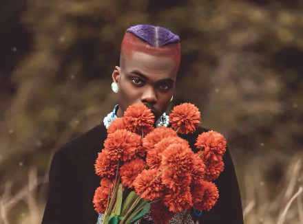 A vibrant HD desktop wallpaper featuring CKay, holding a bouquet of orange flowers, with a striking look and colorful hairstyle against a blurred natural background.