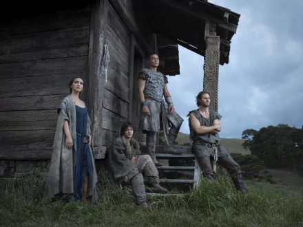 TV show scene from The Lord of the Rings: The Rings of Power — four characters by a rustic wooden hut on a grassy hillside under a cloudy sky. 2K Quad HD PC desktop wallpaper/background.