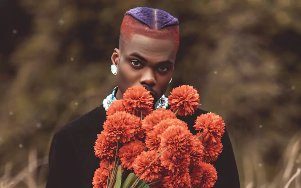 A vibrant HD desktop wallpaper featuring CKay, holding a bouquet of orange flowers, with a striking look and colorful hairstyle against a blurred natural background.