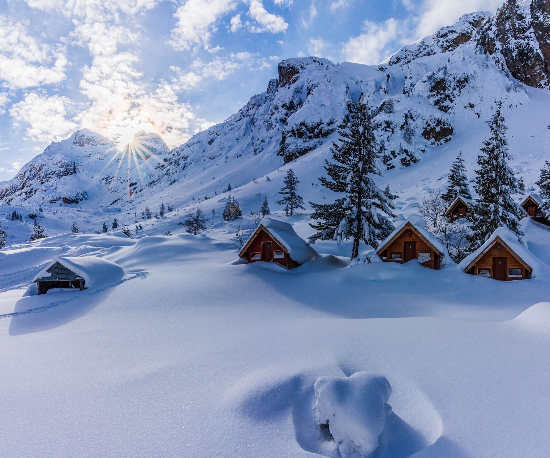 HD desktop wallpaper of a serene winter landscape in Bulgaria, featuring snow-covered wooden cabins and tall pine trees against a bright sky and rugged mountain backdrop.