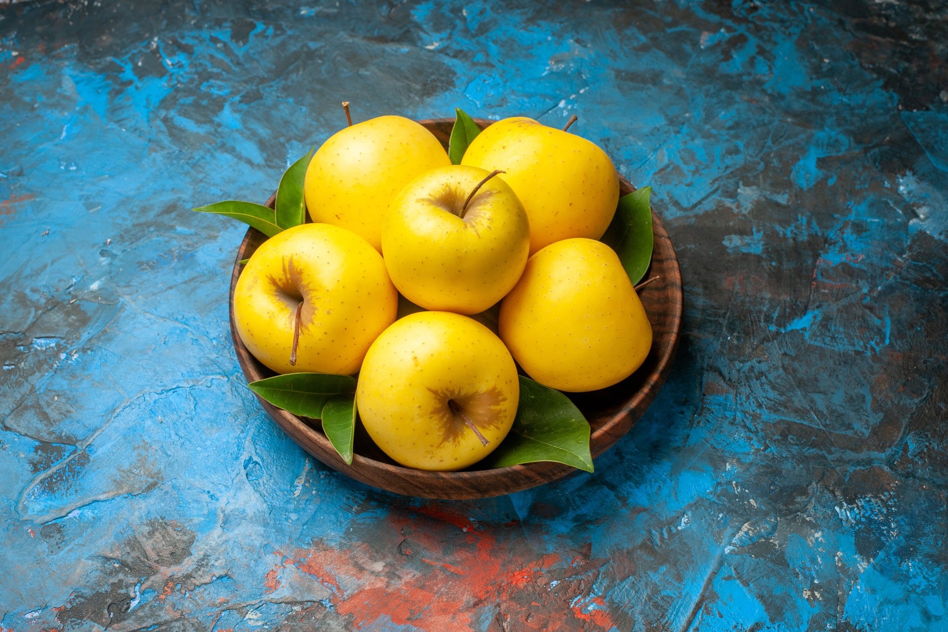5K Ultra HD PC desktop wallpaper: bowl of golden apples with green leaves on a textured blue surface, food photography.