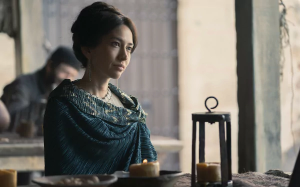 House of the Dragon TV show scene: contemplative woman in a teal embroidered cloak seated at a candlelit table in a moody stone room — 5K Ultra HD PC desktop wallpaper/background.