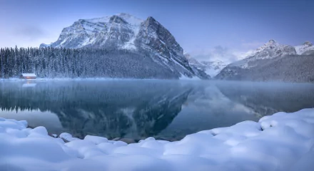 Winter view of Lake Louise in Banff National Park, Canada, showcasing snow-covered shores and crystal-clear reflection of the mountains in the calm lake waters.