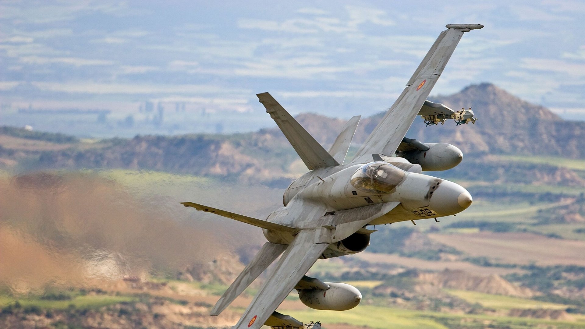 Military McDonnell Douglas F/A-18 Hornet flying low over a mountainous landscape, captured in an HD PC desktop wallpaper background.