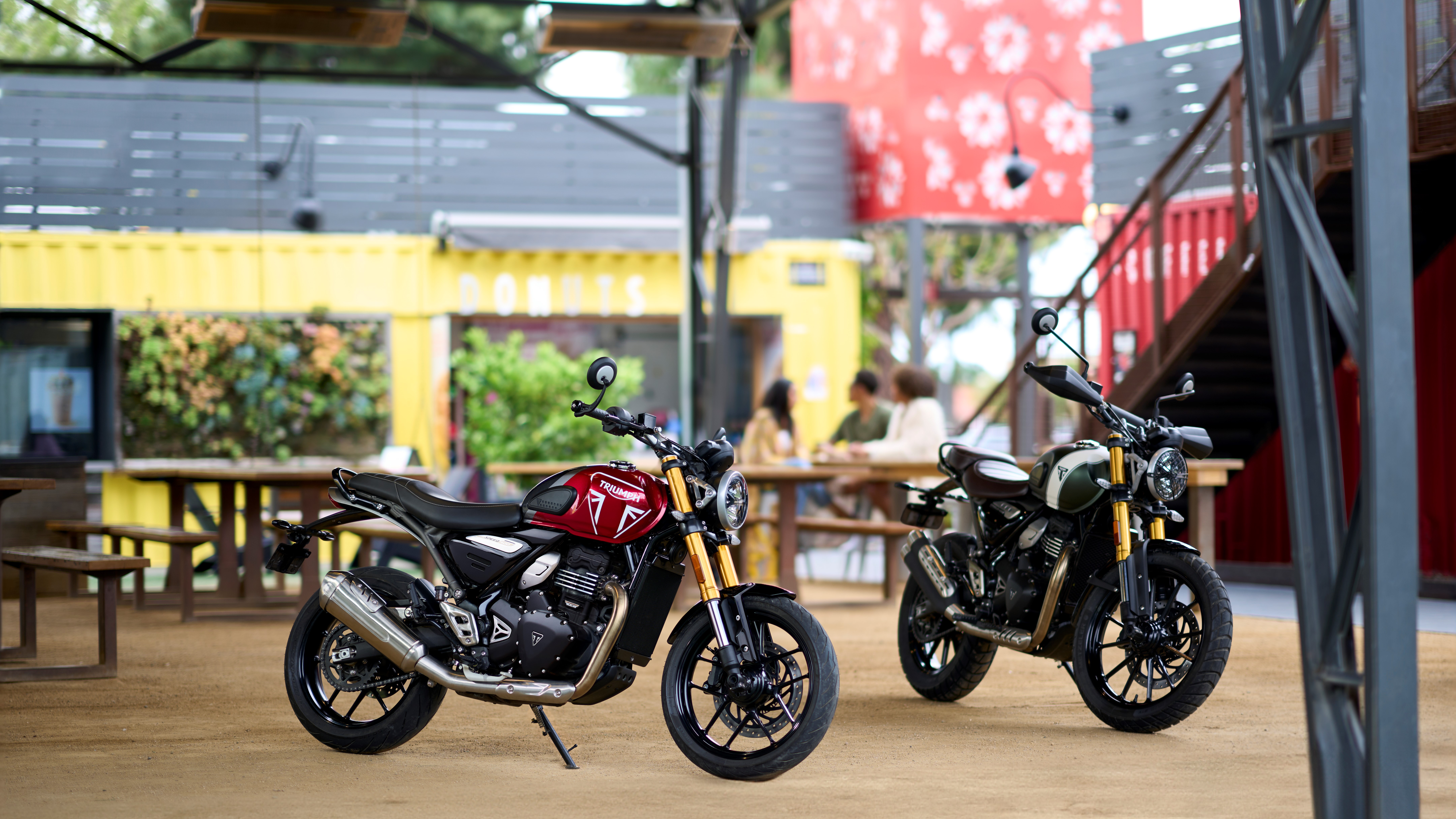 HD wallpaper featuring Triumph Speed 400 and Triumph Scrambler 400 X motorcycles parked in an urban outdoor cafe setting.