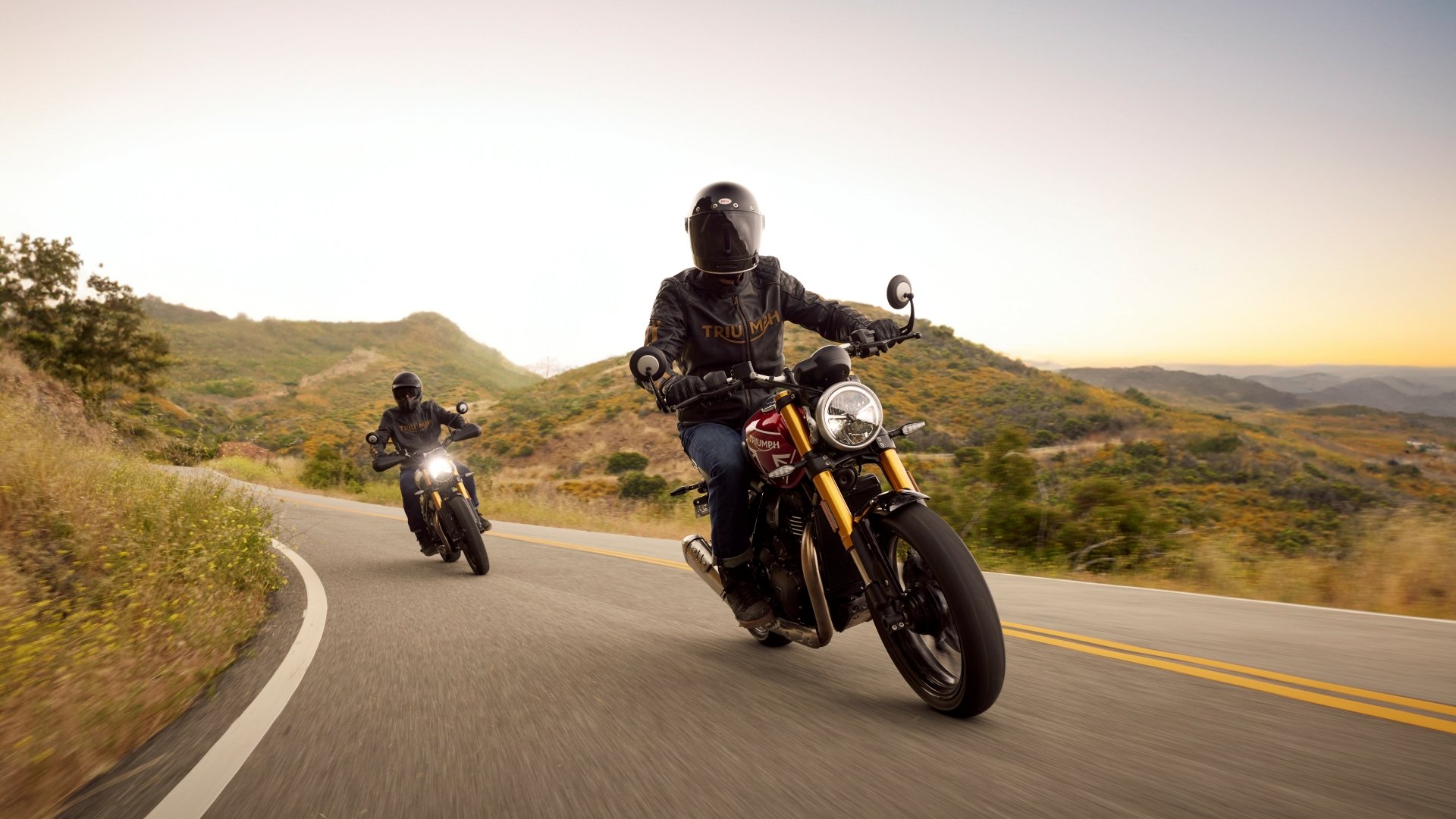 A thrilling HD desktop wallpaper featuring two riders on Triumph Speed 400 motorcycles navigating a winding road through scenic hills at sunset.