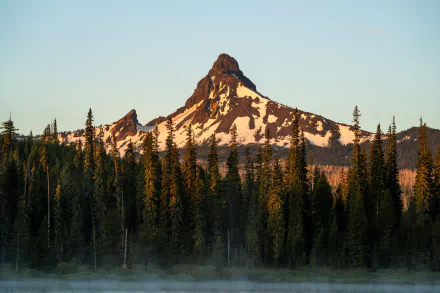 HD desktop wallpaper showcasing a snow-capped mountain peak rising above a dense forest under a clear sky, embodying the beauty of nature and mountainous landscapes.