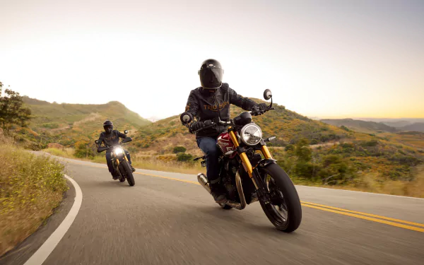 A thrilling HD desktop wallpaper featuring two riders on Triumph Speed 400 motorcycles navigating a winding road through scenic hills at sunset.