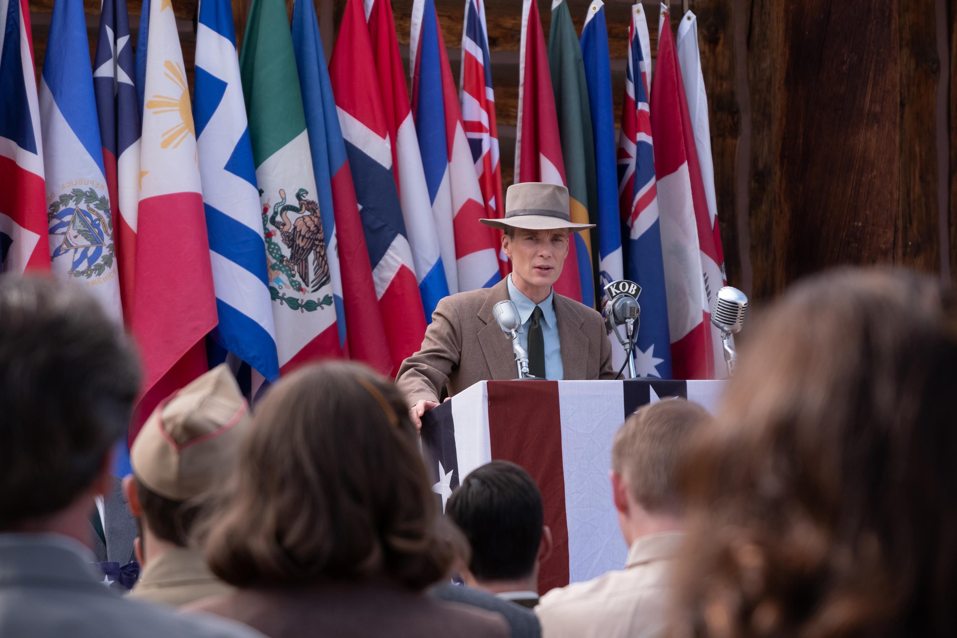 HD Wallpaper of a man resembling Cillian Murphy as Oppenheimer speaking at a podium surrounded by international flags, ideal for desktop background.
