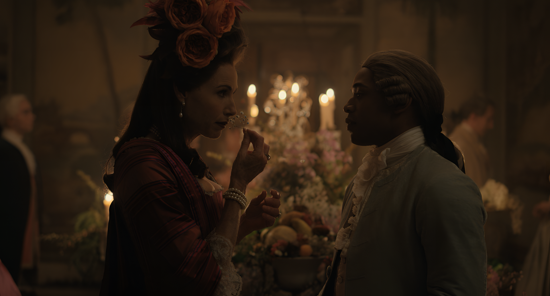 HD desktop wallpaper featuring an elegant scene with two people in historical Chevalier-style costumes, conversing intimately in a candlelit room adorned with flowers and a chandelier.