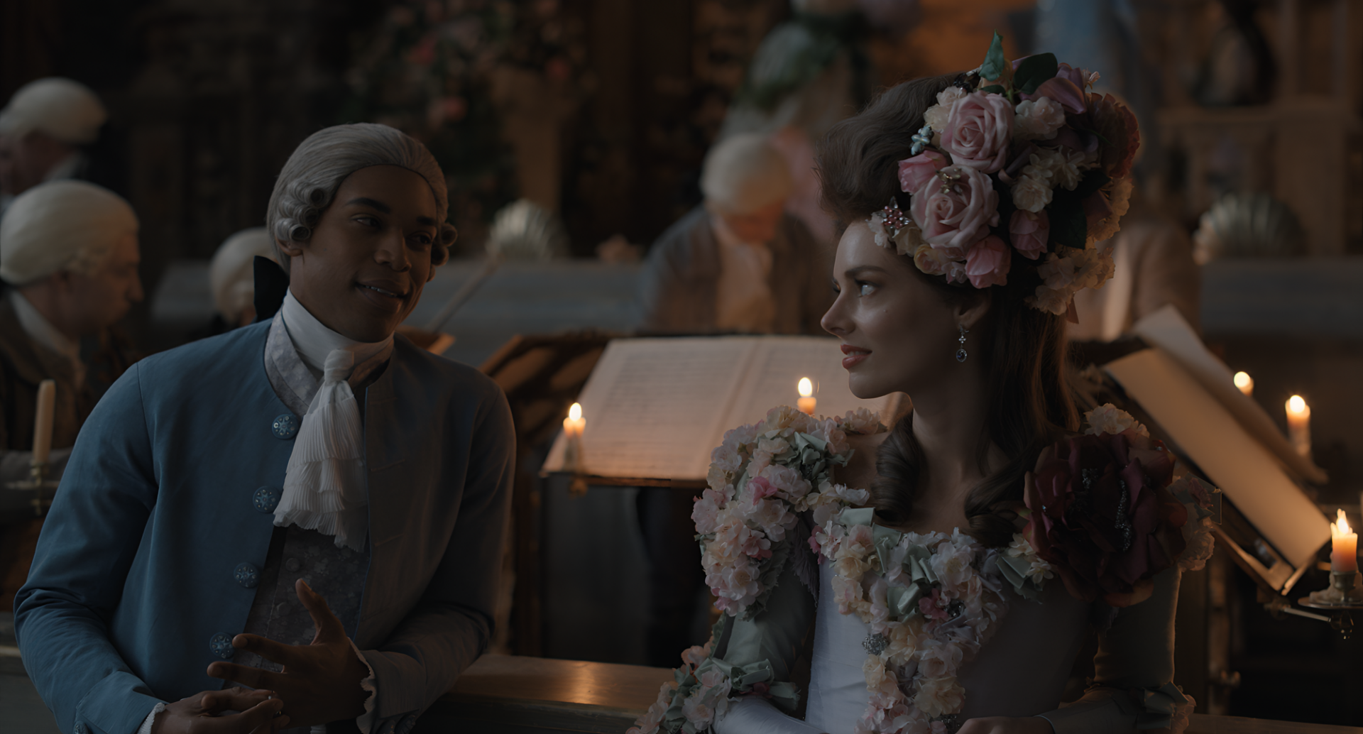 HD wallpaper of a chevalier-themed scene with two elegantly dressed individuals, one wearing a floral headdress, in a candlelit historical setting.