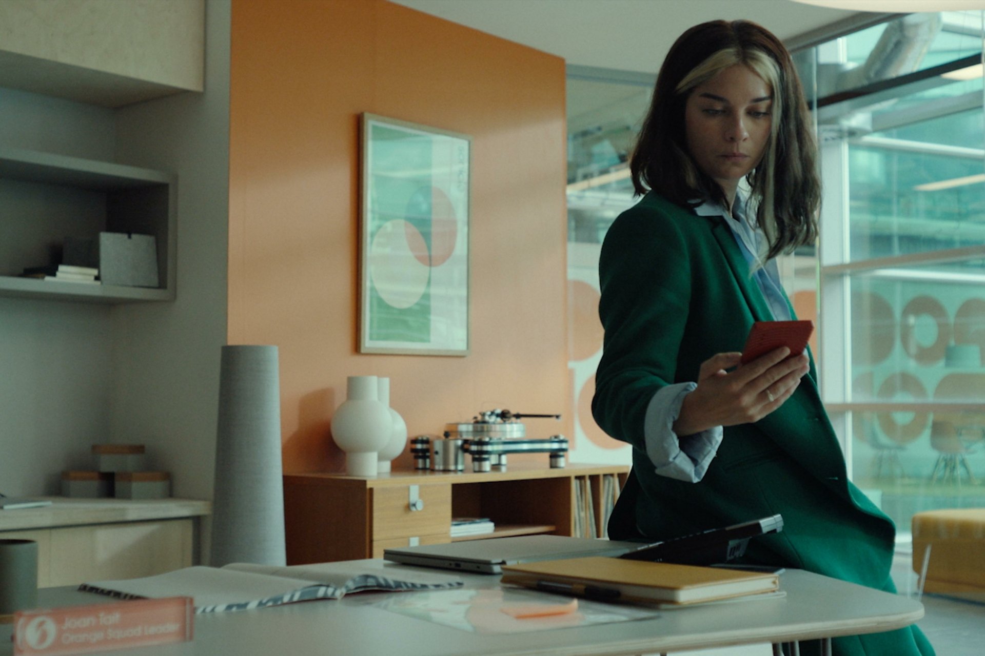Alt Text: HD wallpaper featuring a scene from Black Mirror with actress Annie Murphy wearing a green coat, focused on her phone in a modern office setting with warm pastel tones.