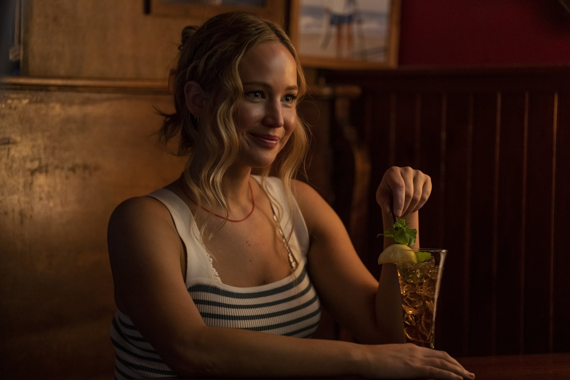 A scene from the movie No Hard Feelings, featuring a woman with wavy hair sitting at a bar, smiling as she stirs a drink in a warm, inviting atmosphere.