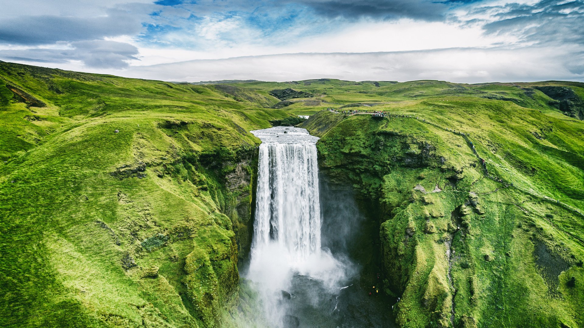 A stunning 4K Ultra HD nature wallpaper featuring Skógafoss waterfall cascading between lush green cliffs under a cloudy sky.