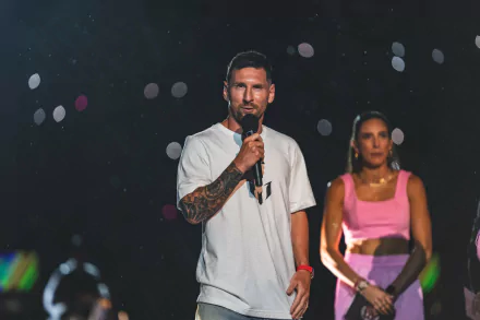 HD desktop wallpaper featuring Lionel Messi speaking into a microphone on stage, with a woman in the background and colorful bokeh lights.