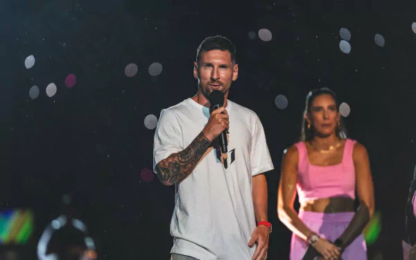 HD desktop wallpaper featuring Lionel Messi speaking into a microphone on stage, with a woman in the background and colorful bokeh lights.