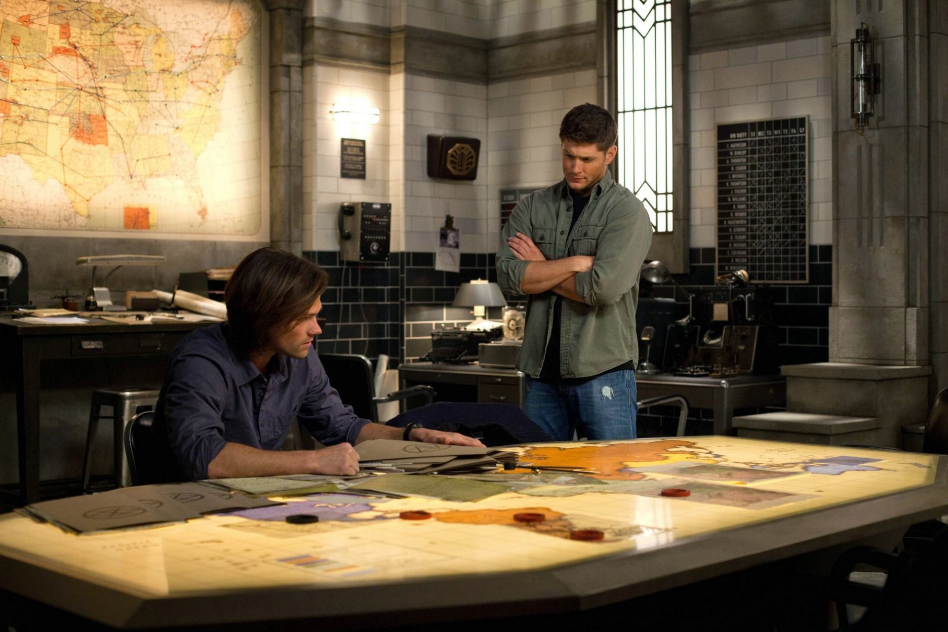 HD desktop wallpaper of two men in a dim investigation room from the TV show Supernatural: one seated at a map-covered table, the other standing with arms crossed.