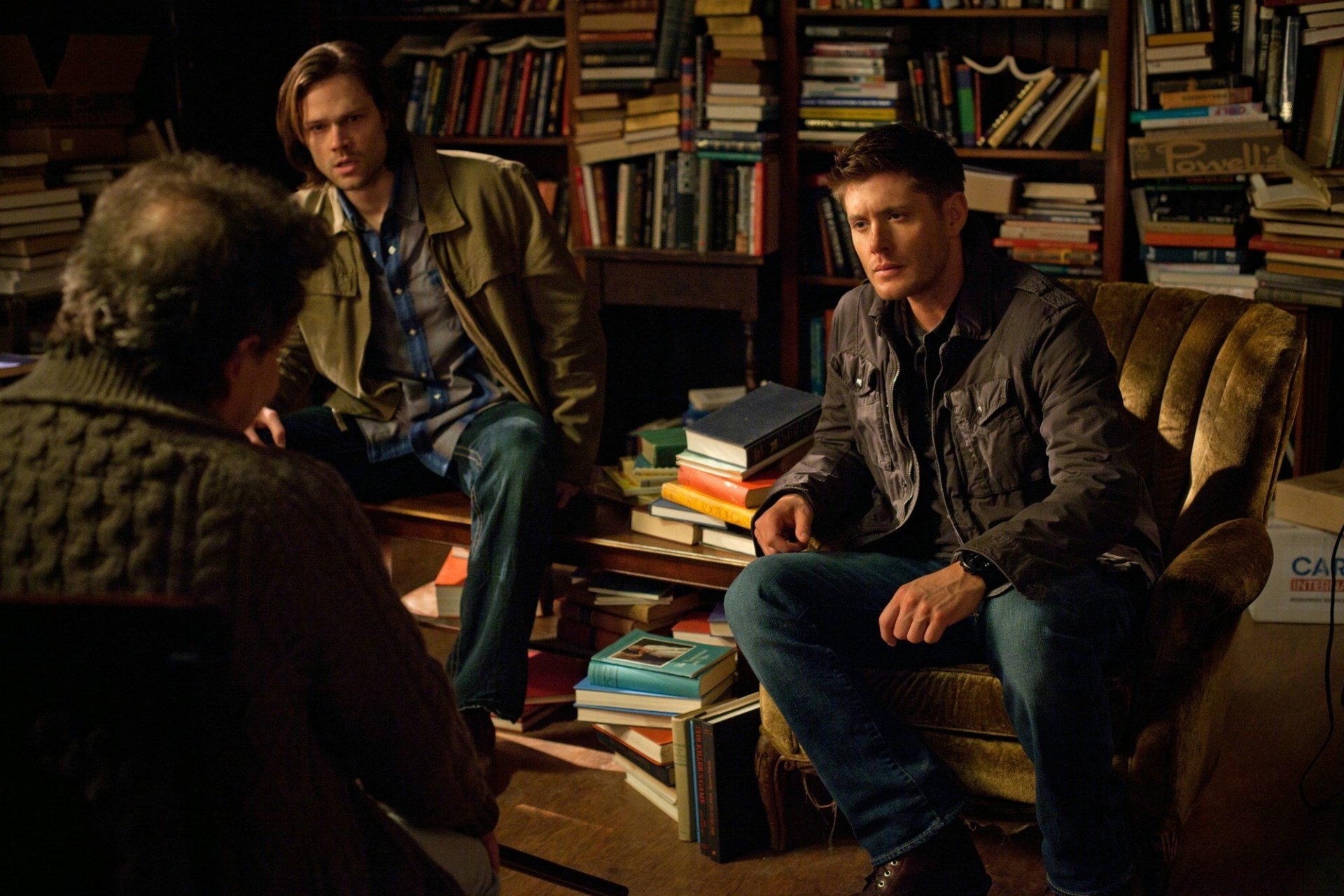 HD desktop wallpaper: Dean and Sam Winchester from Supernatural sit in a cluttered study surrounded by stacks of books, engaged in a tense conversation.