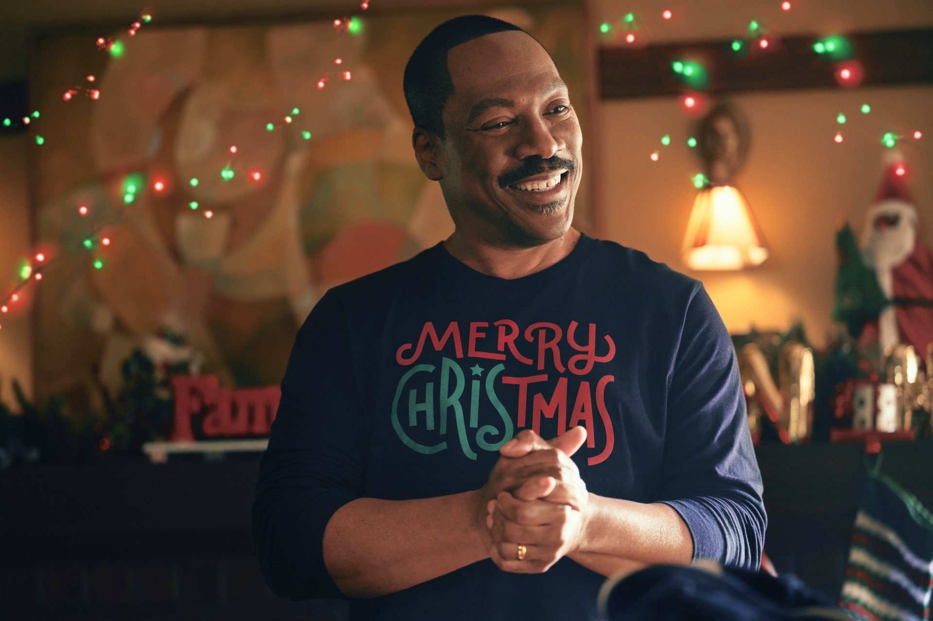 Man smiling in a festive room decorated with Christmas lights and holiday decor, wearing a 'Merry Christmas' sweater, from the movie 'Candy Cane Lane' - HD desktop wallpaper background.