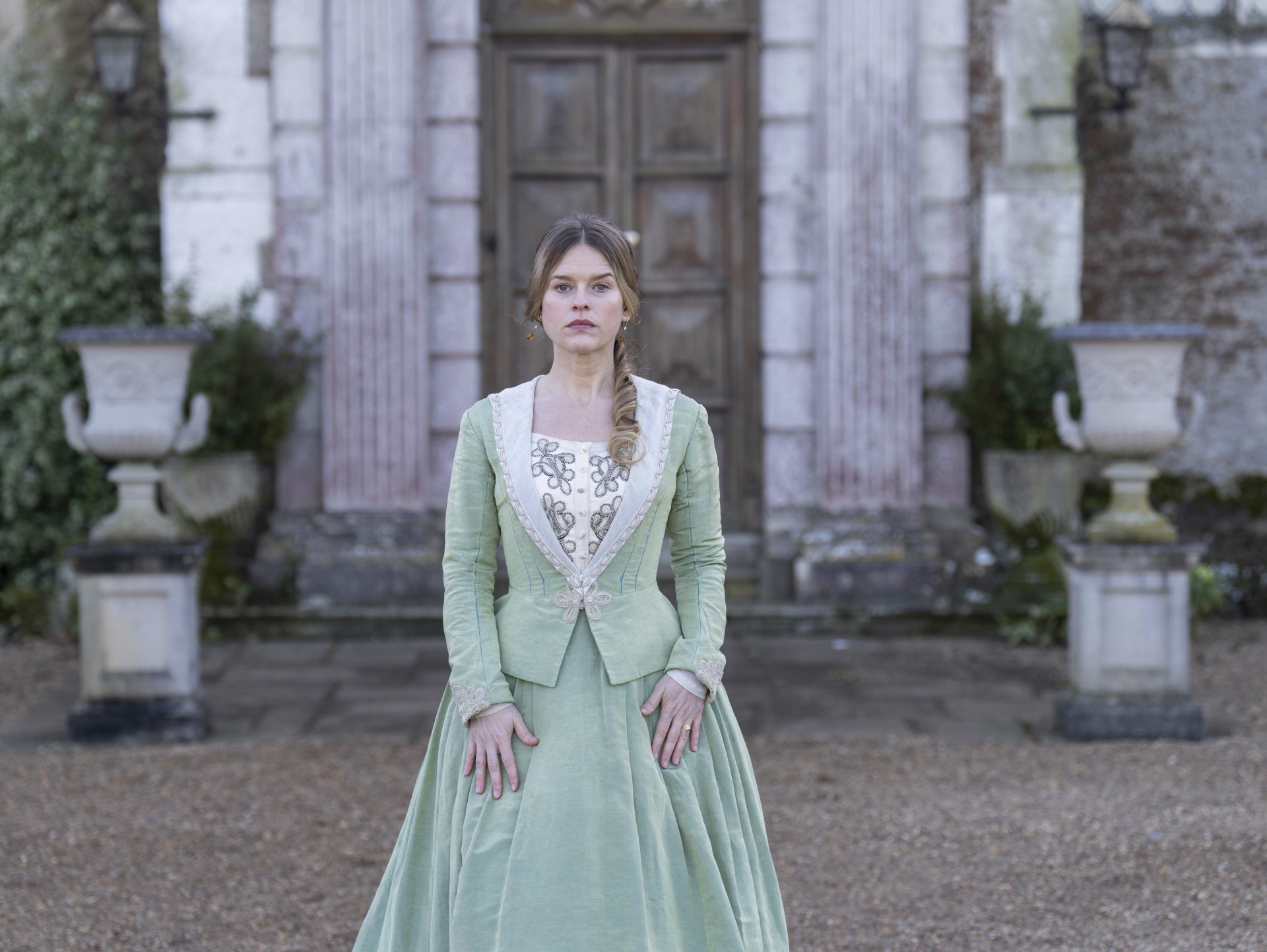 A captivating HD wallpaper from the TV show Belgravia: The Next Chapter, featuring a young woman in a green gown standing in front of an elegant historic backdrop.