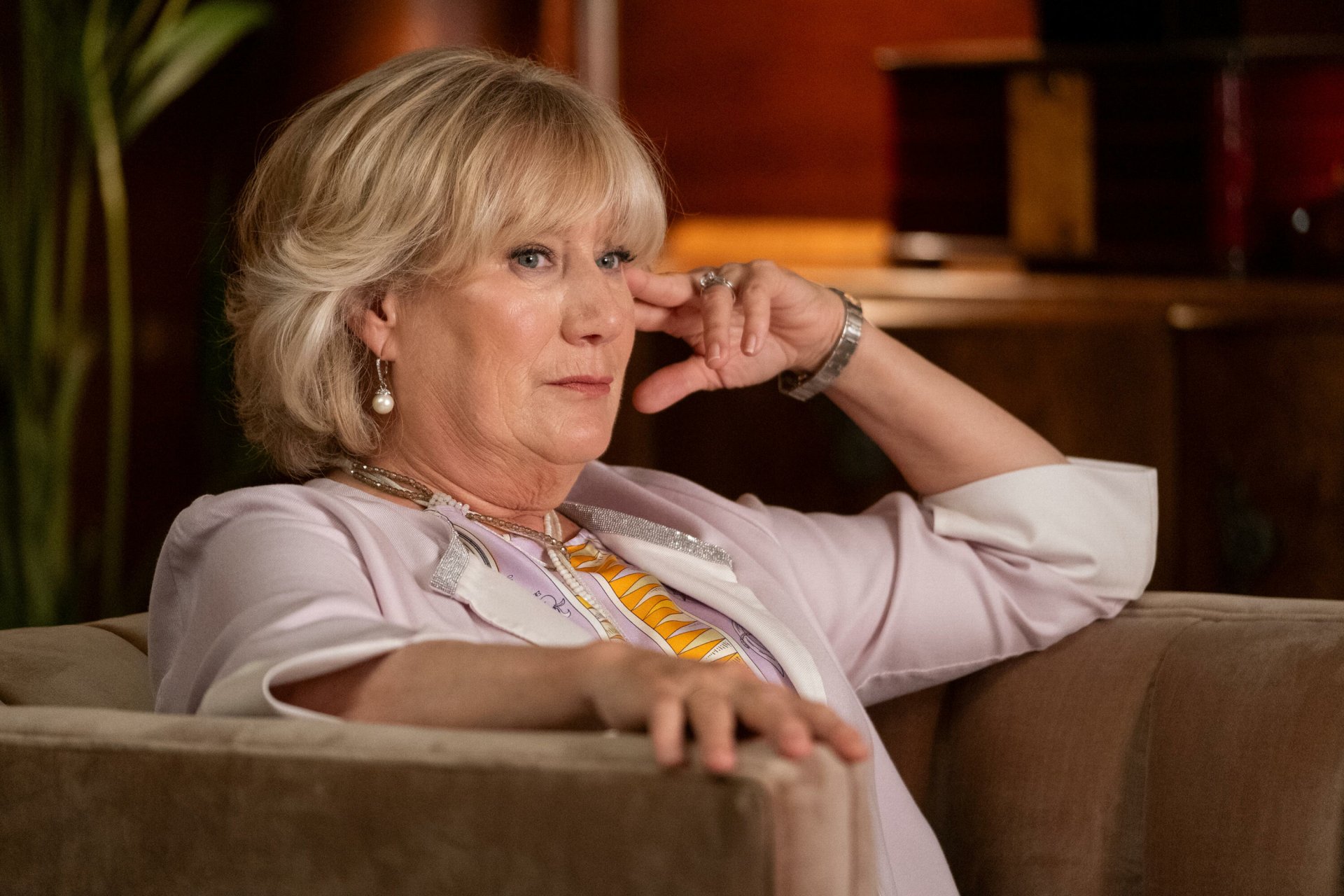 A thoughtful character from the TV show Death and Other Details sits on a couch, deep in contemplation, surrounded by a warm, inviting background.