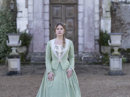 A captivating HD wallpaper from the TV show Belgravia: The Next Chapter, featuring a young woman in a green gown standing in front of an elegant historic backdrop.
