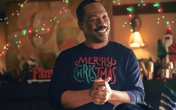Man smiling in a festive room decorated with Christmas lights and holiday decor, wearing a 'Merry Christmas' sweater, from the movie 'Candy Cane Lane' - HD desktop wallpaper background.