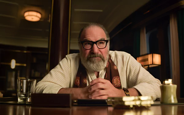 HD wallpaper featuring a contemplative man (actor from a TV show) sitting at a table, possibly portraying a character involved with death and other details, in a dimly-lit room with vintage decor.