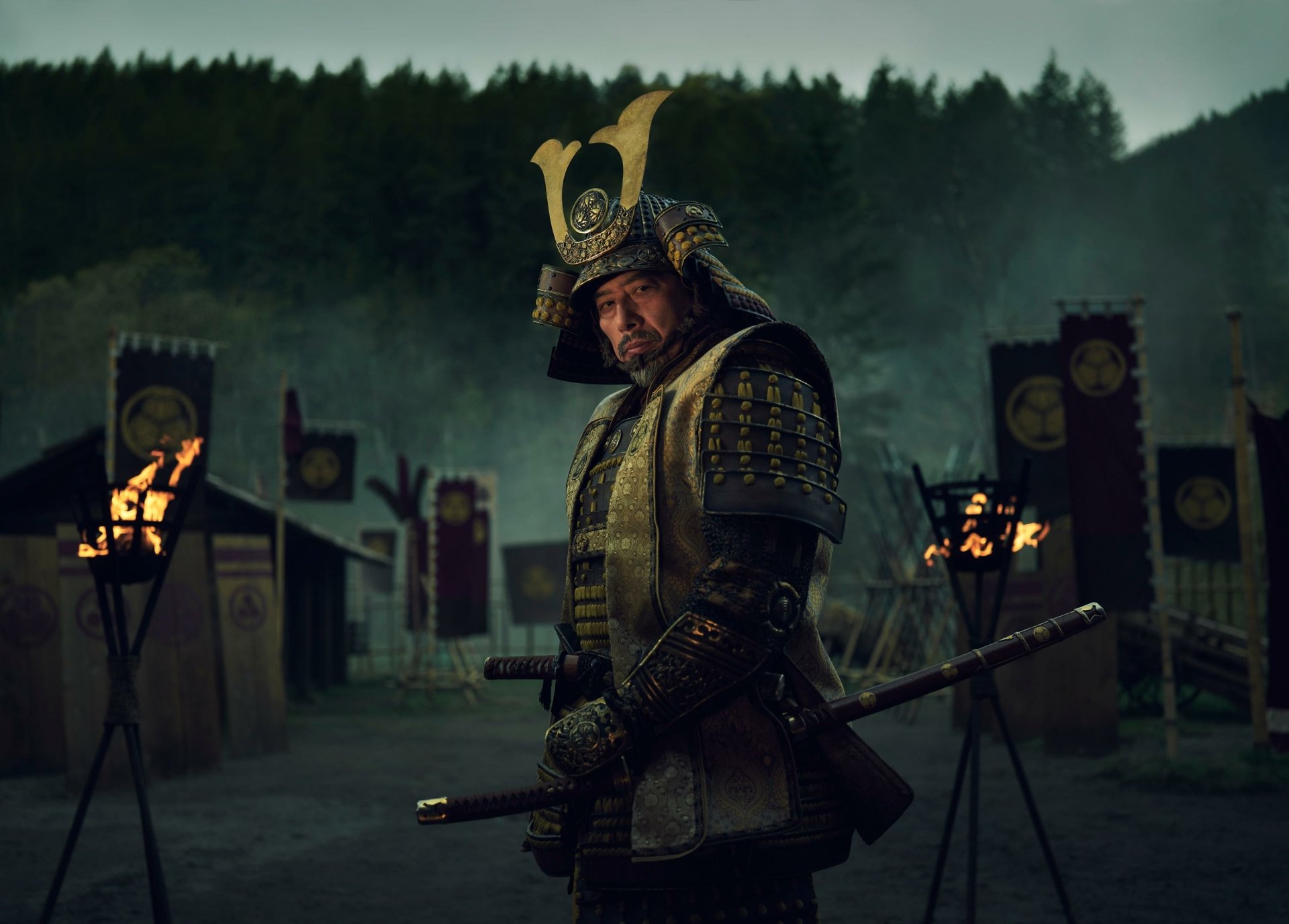 HD Wallpaper featuring a samurai from the 2024 TV show Shogun, dressed in traditional armor, standing solemnly in a dusk-lit encampment with banners and torches in the background.