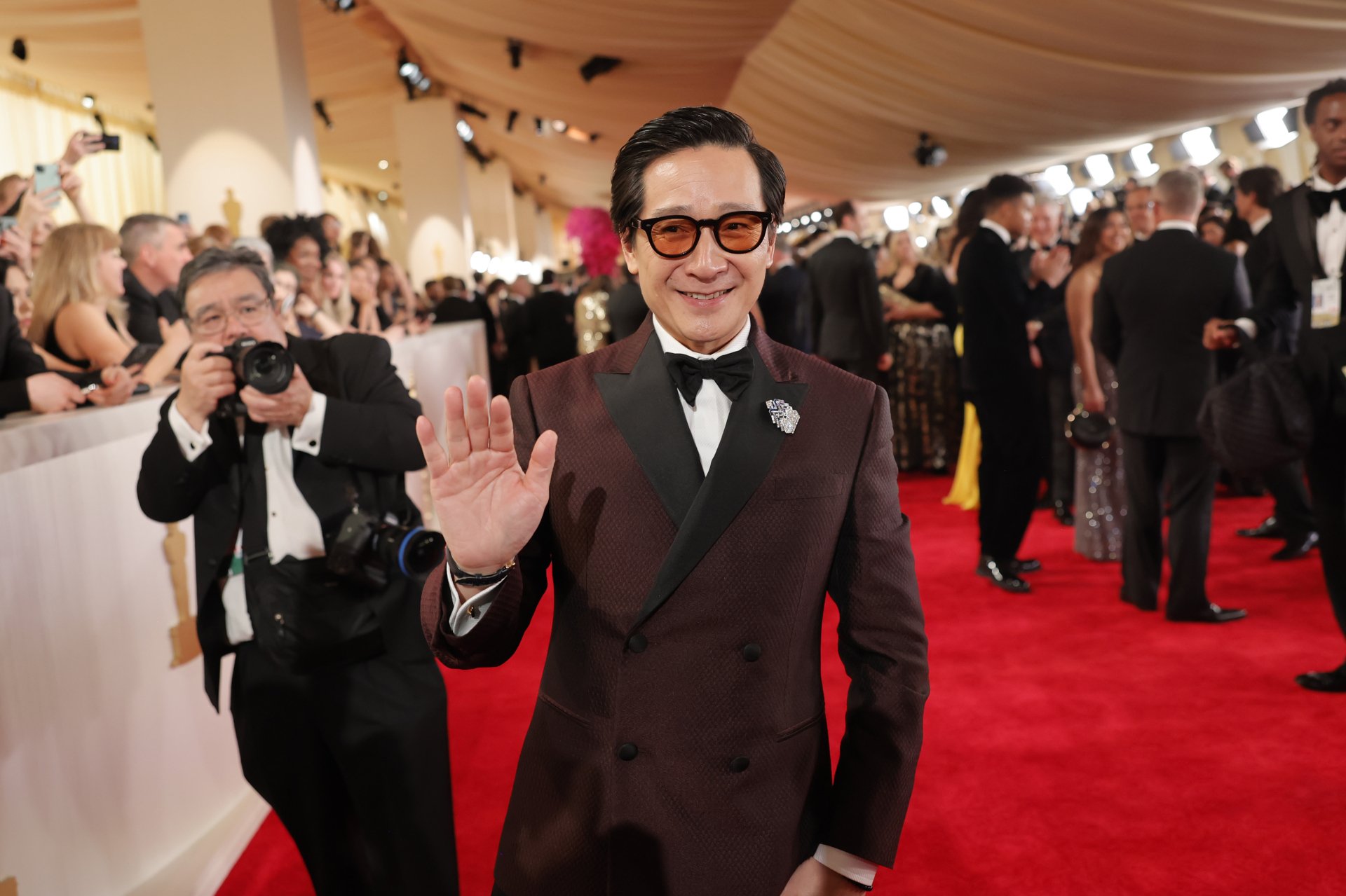 Ke Huy Quan smiling and waving on the red carpet at the Academy Awards, suited for a HD desktop wallpaper.