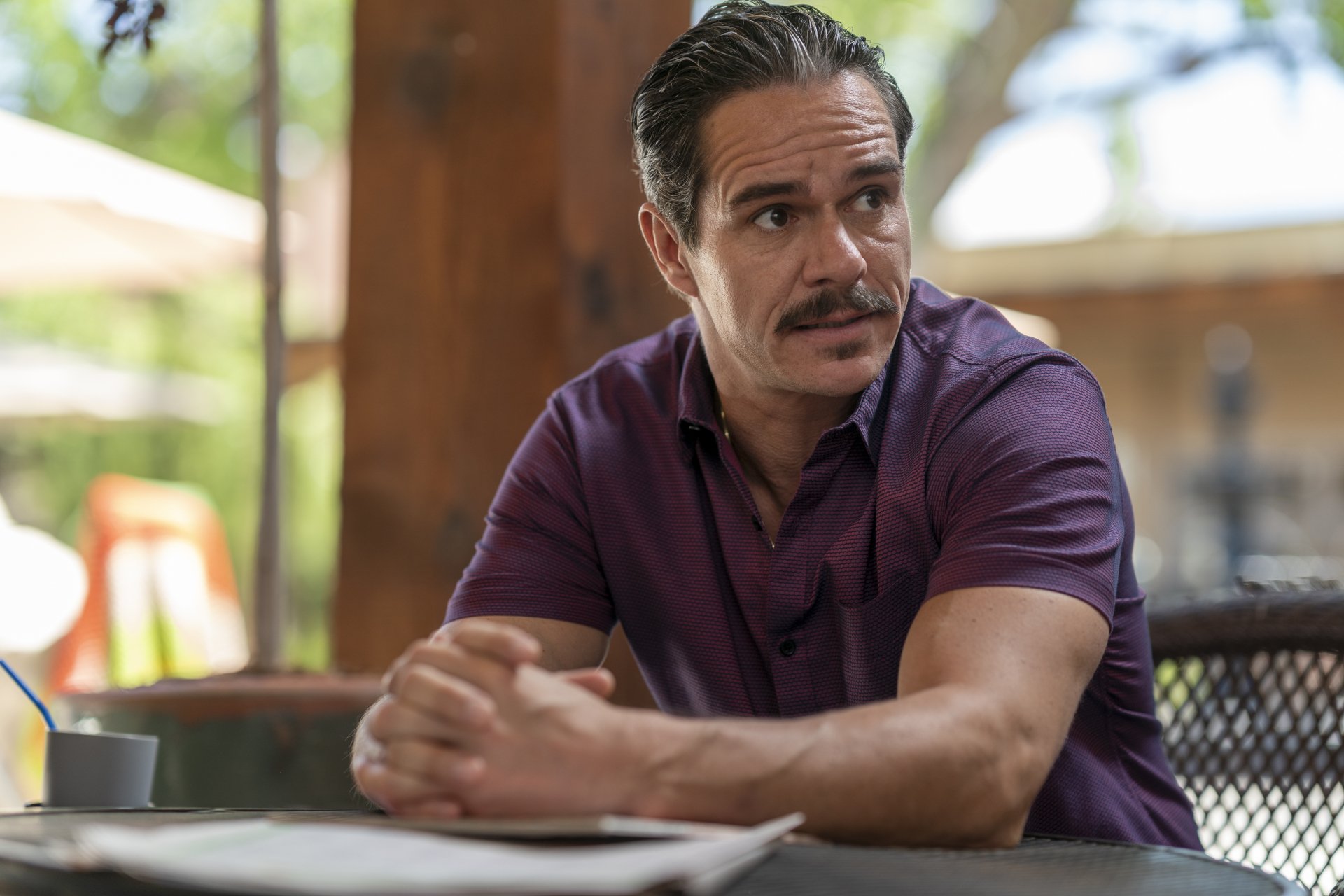 A still from the TV show Better Call Saul, featuring a man with a mustache sitting at a table, deep in thought, set against a soft outdoor background. HD desktop wallpaper quality.