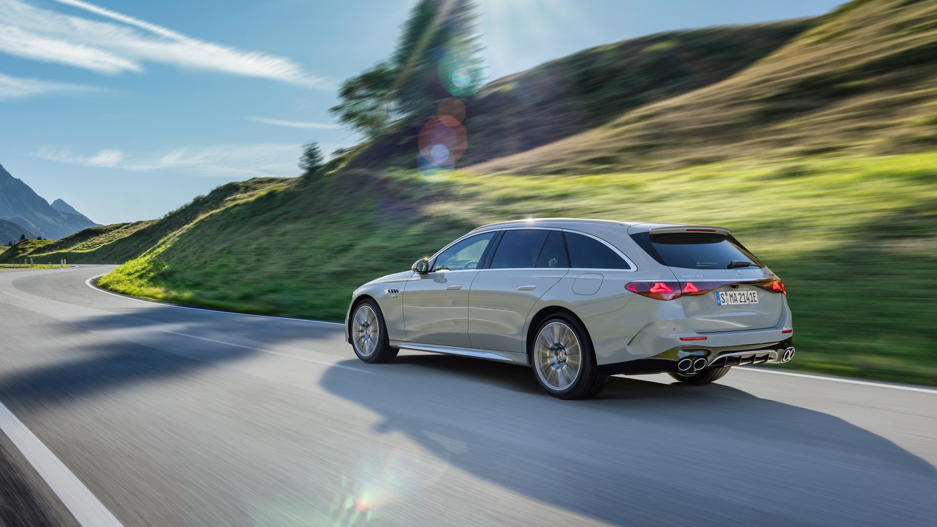A sleek Mercedes-AMG E53 Hybrid cruises along a winding road, surrounded by lush green hills and blue skies, captured in this stunning HD desktop wallpaper.