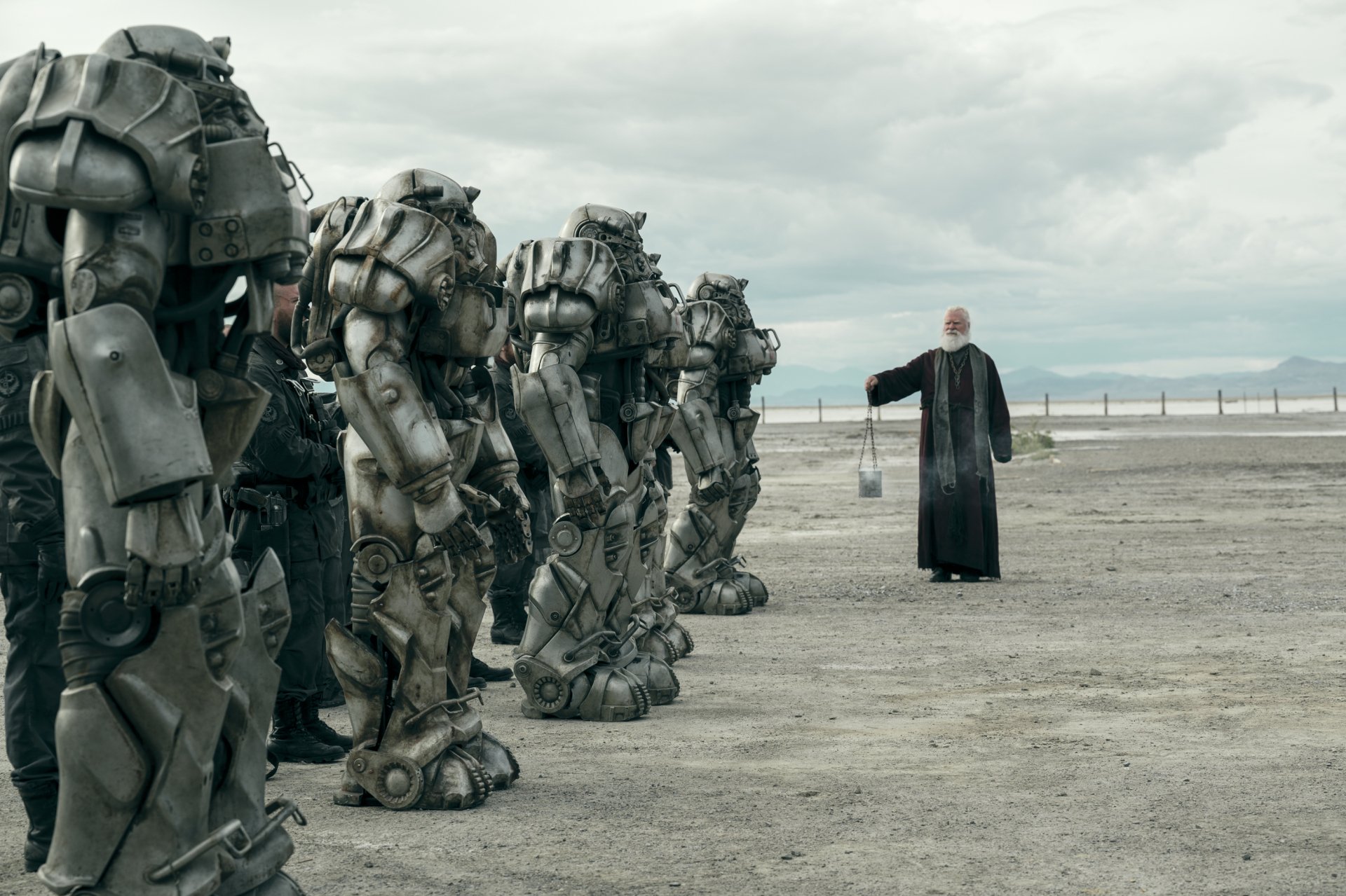 A scene from the TV show Fallout featuring a line of characters in heavy power armor standing alongside a solitary figure in a long coat, set in a desolate landscape, providing a dramatic backdrop for a desktop wallpaper.