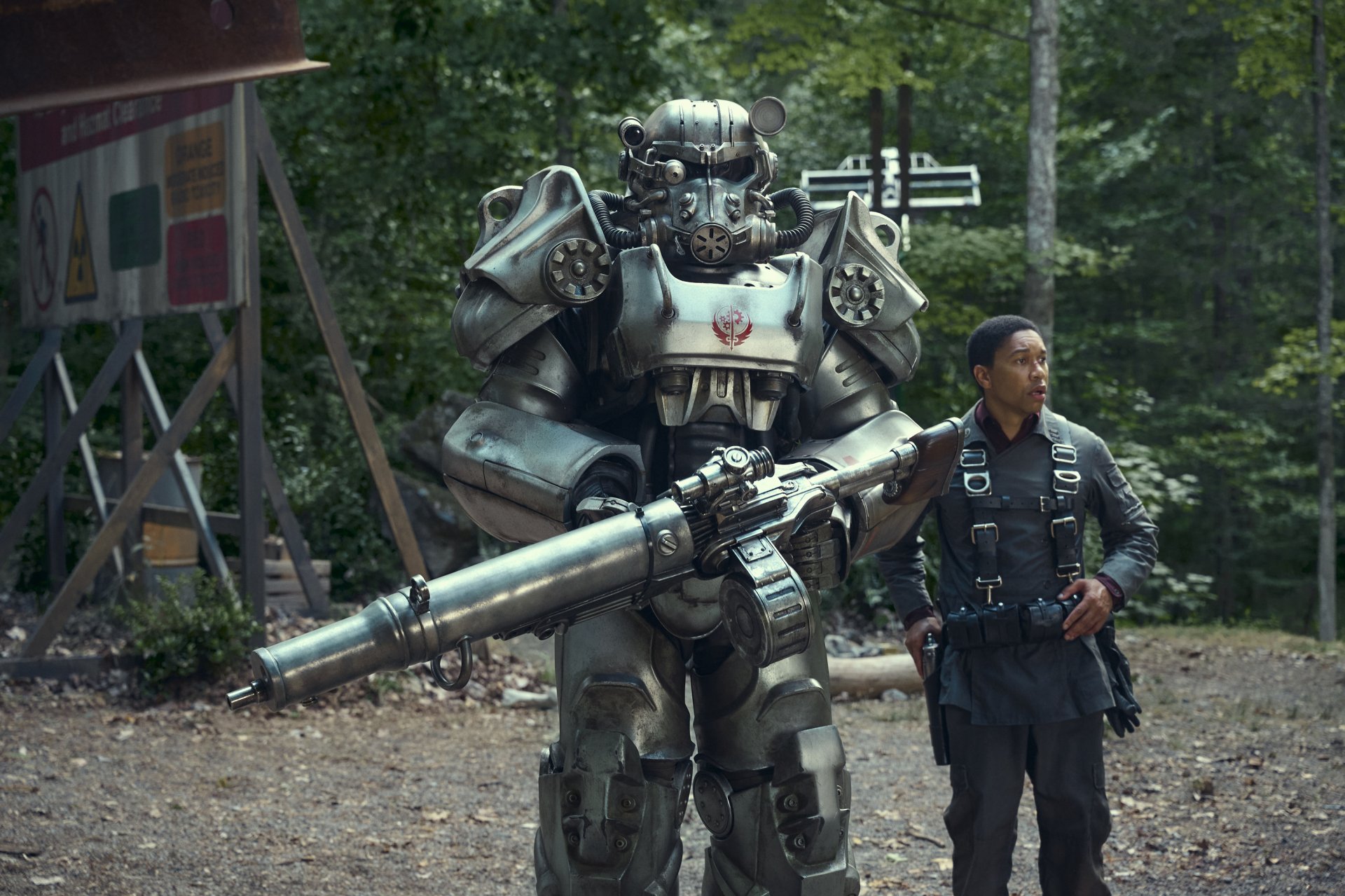 HD desktop wallpaper featuring a scene from the TV show Fallout with a character in heavy power armor standing next to another character in combat gear against a forested backdrop.