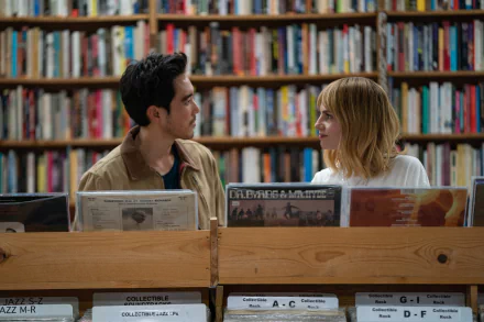 A scene from The Greatest Hits featuring Lucy Boynton and Justin H. Min, set in a cozy record shop surrounded by shelves of books. A captivating moment captured in HD.