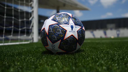 High-definition desktop wallpaper featuring a UEFA Champions League soccer ball on a lush green pitch with a goalpost in the background, under a clear blue sky.