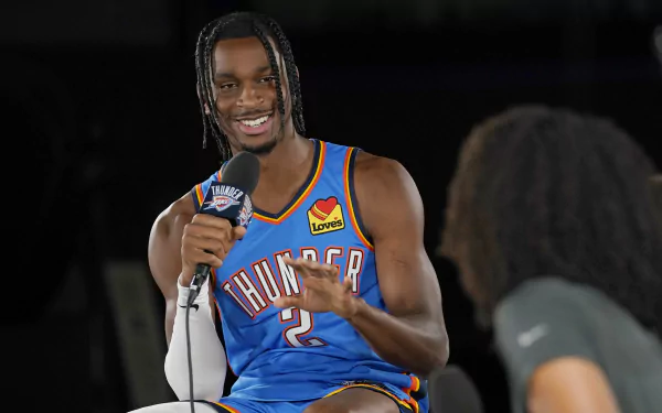 Shai Gilgeous-Alexander of the Oklahoma City Thunder smiles while speaking into a microphone, showcasing his dynamic presence in this vibrant HD background.