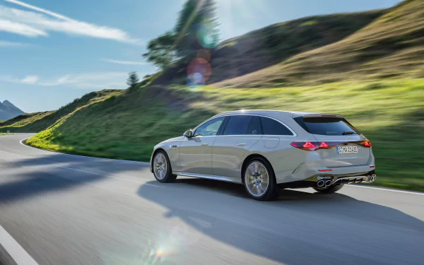 A sleek Mercedes-AMG E53 Hybrid cruises along a winding road, surrounded by lush green hills and blue skies, captured in this stunning HD desktop wallpaper.