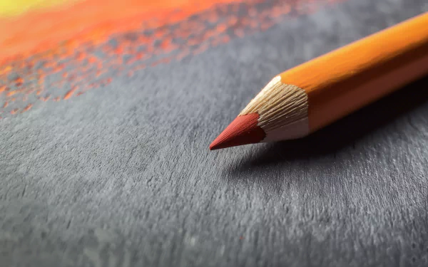 Close-up HD image of a sharpened orange colored pencil with a red tip, lying on a textured surface with a gradient of red hues in the background. Perfect for desktop wallpaper or background.