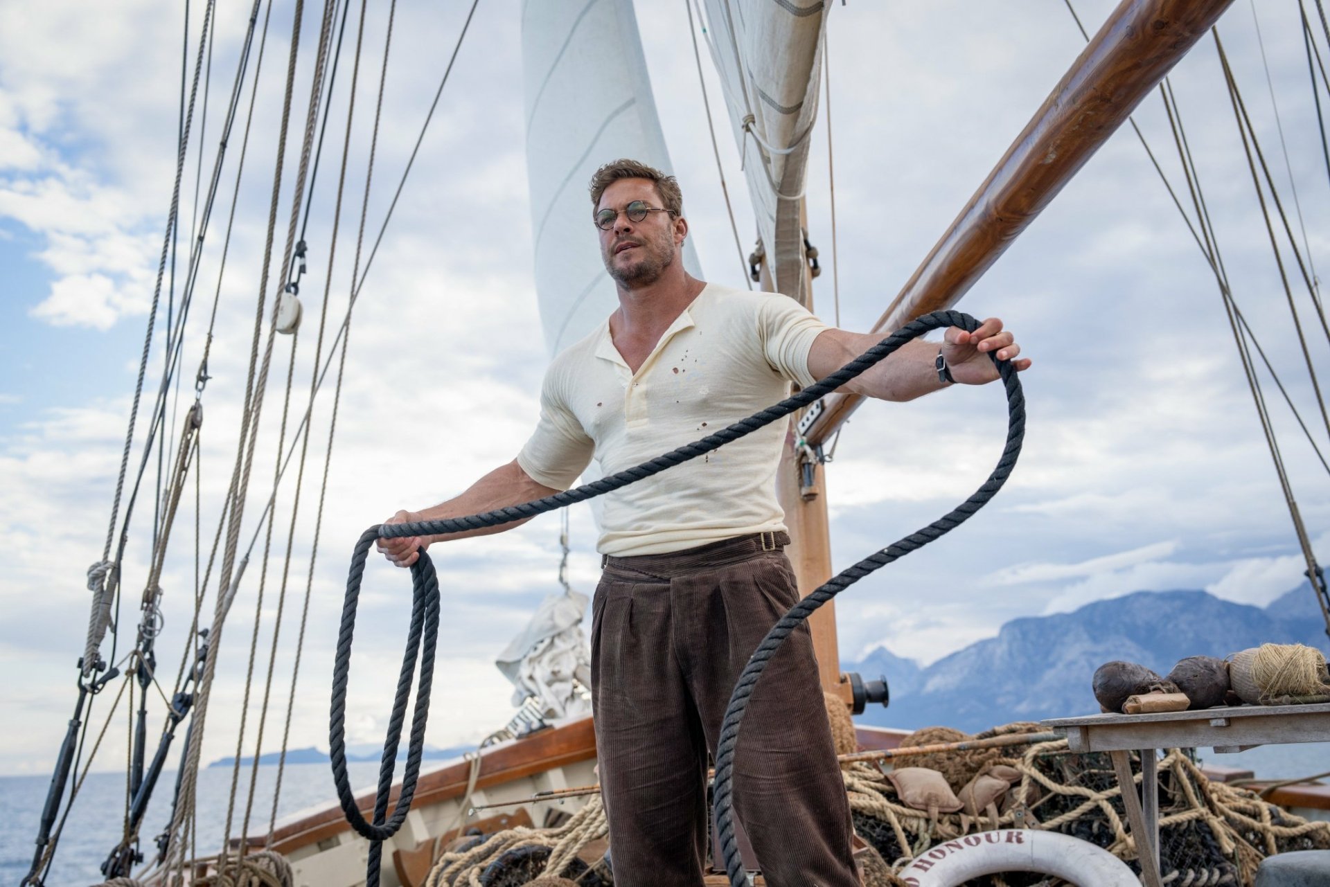 A man stands aboard a sailing ship holding thick ropes, with clouds and mountains in the background. This HD image is styled as a desktop wallpaper, associated with the movie The Ministry of Ungentlemanly Warfare.