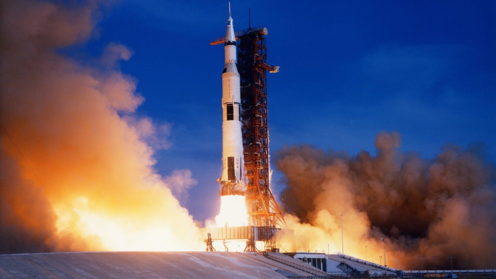 HD wallpaper of the Saturn V rocket launching, enveloped in smoke and flames, set against a clear sky.