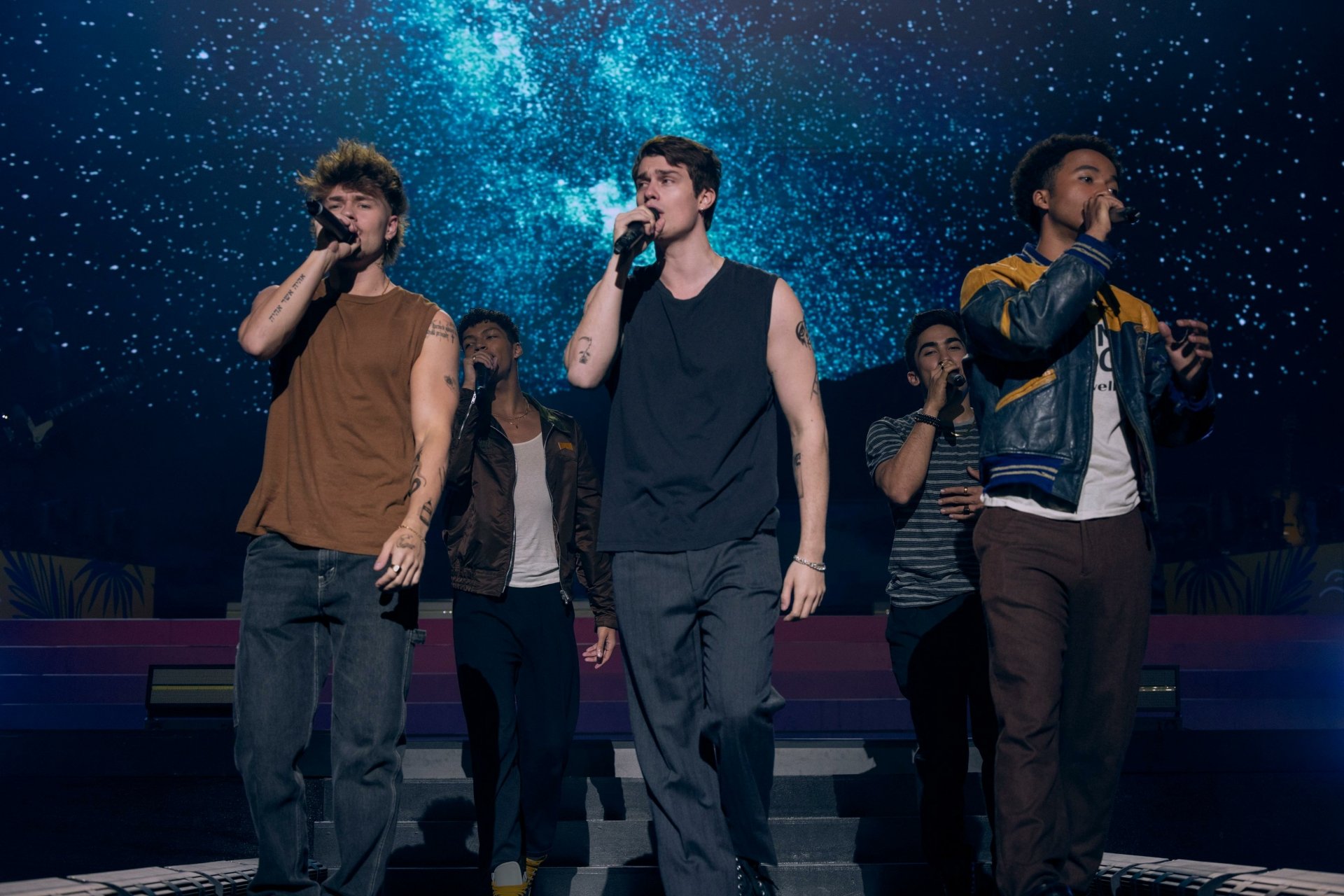 HD wallpaper from The Idea of You featuring Nicholas Galitzine performing on stage with a group under a cosmic blue backdrop.