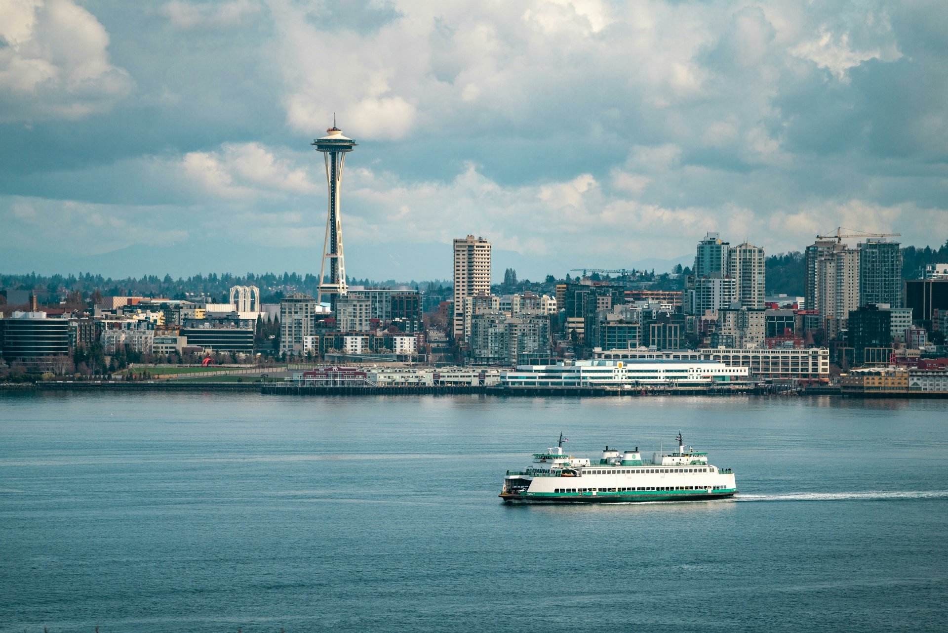 A high-definition desktop wallpaper featuring a ferry sailing on the water with Seattle's skyline and the Space Needle in the background under a partly cloudy sky.