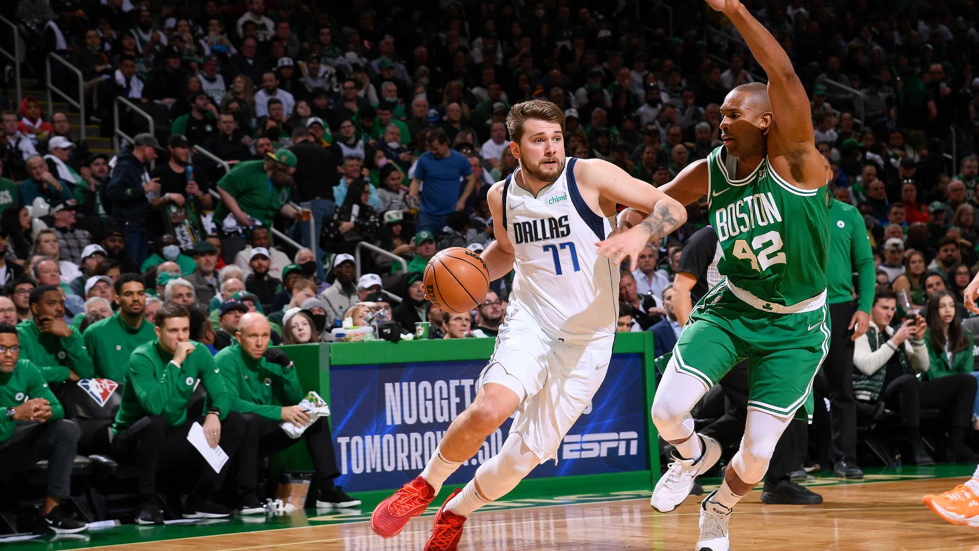 Luka Dončić of the Dallas Mavericks dribbles past a Boston Celtics defender during an NBA game. The crowded arena captures the intense atmosphere, making this a captivating HD desktop wallpaper and background.
