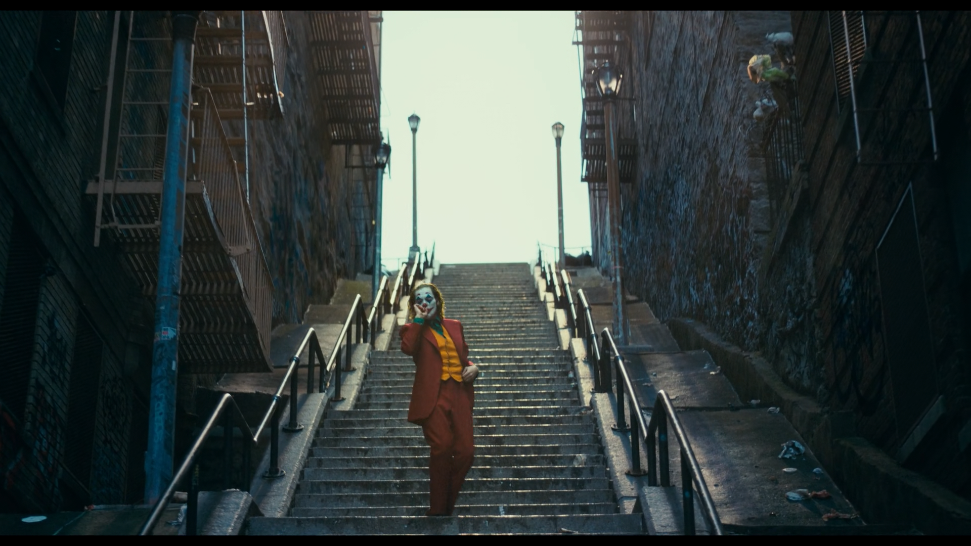 HD PC desktop wallpaper featuring the Joker in his iconic orange suit, walking down a graffiti-covered urban staircase with a bright sky above.
