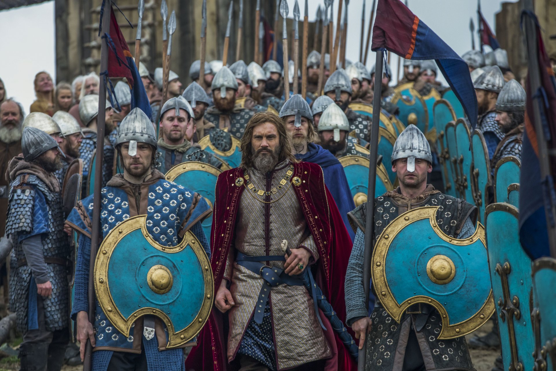 Clive Standen as Rollo from the TV show Vikings leads an army of warriors with blue shields and helmets. This HD desktop wallpaper captures a dramatic scene from the series.