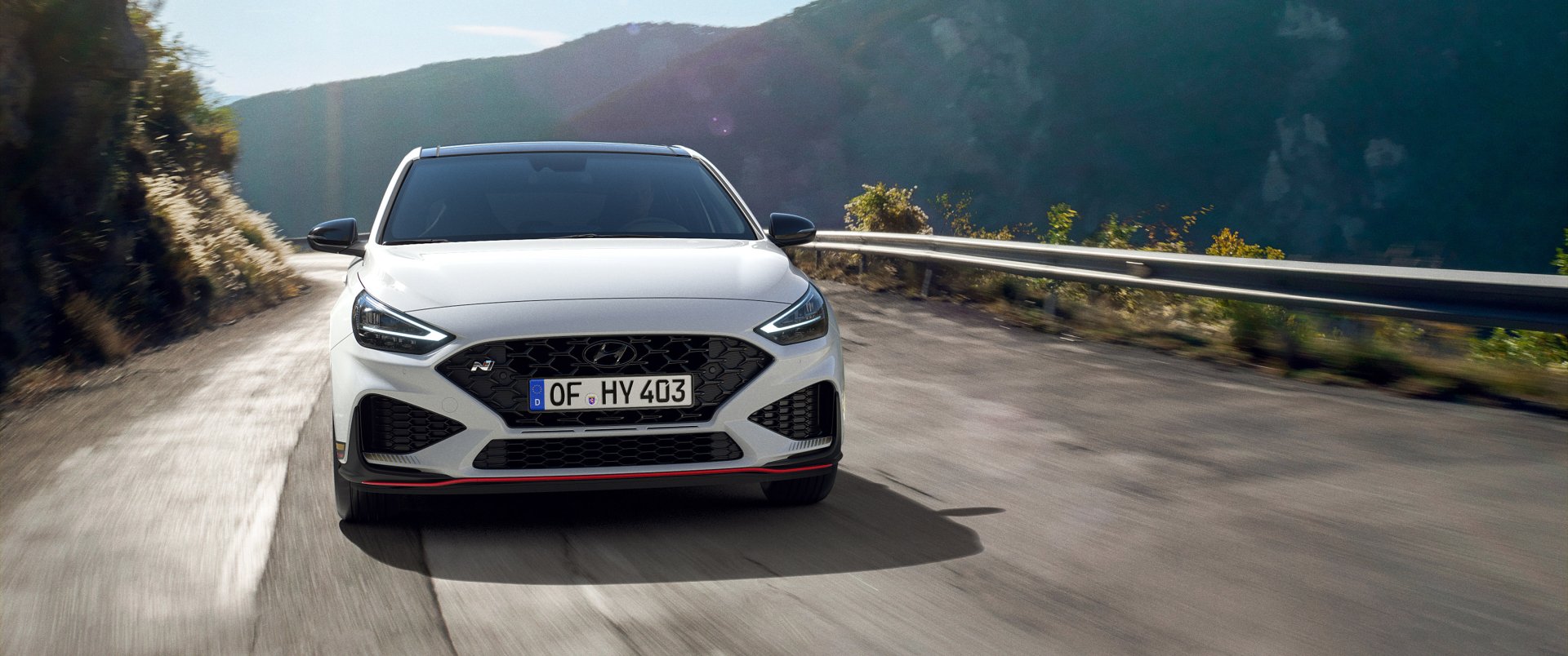 HD wallpaper of a Hyundai i30 car driving on a mountain road, captured from the front with scenic mountainous background. The image emphasizes the sleek design of the vehicle.
