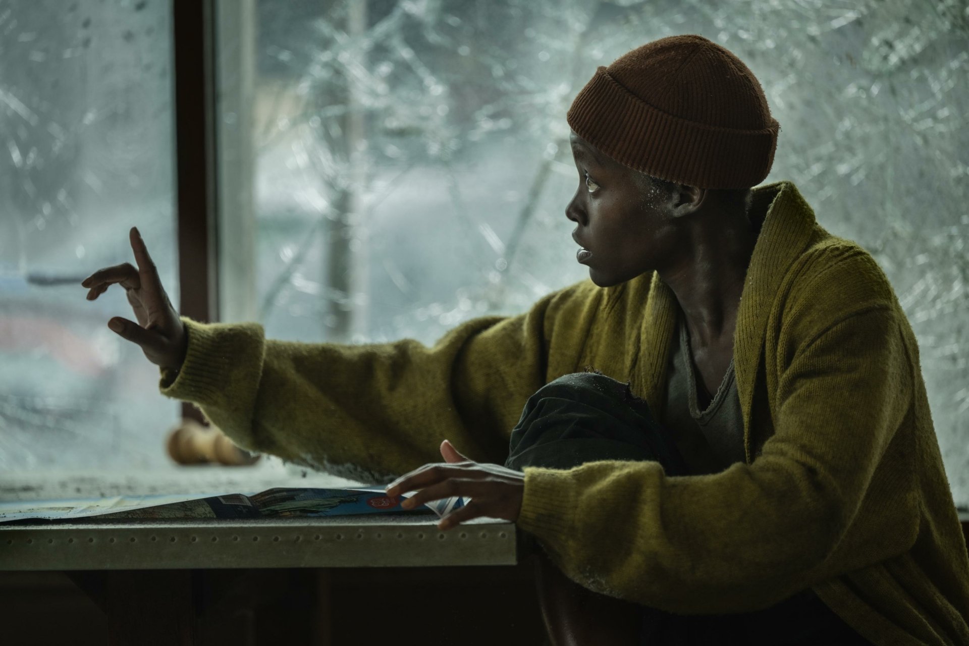 A HD desktop wallpaper from the movie A Quiet Place: Day One, featuring Lupita Nyong'o in a tense scene by a cracked window.